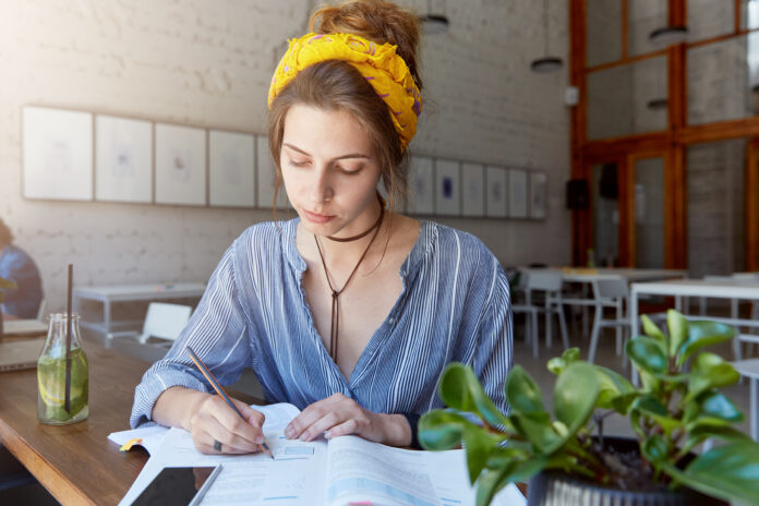 Portrait of scientific worker sitting in cabinet writing down something with serious look. Young educated woman siting in cozy classroom writing and reading books looking down using mobile phone