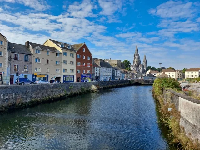 River Lee in Cork with riverside buildings, a bridge and church spires under a blue sky
